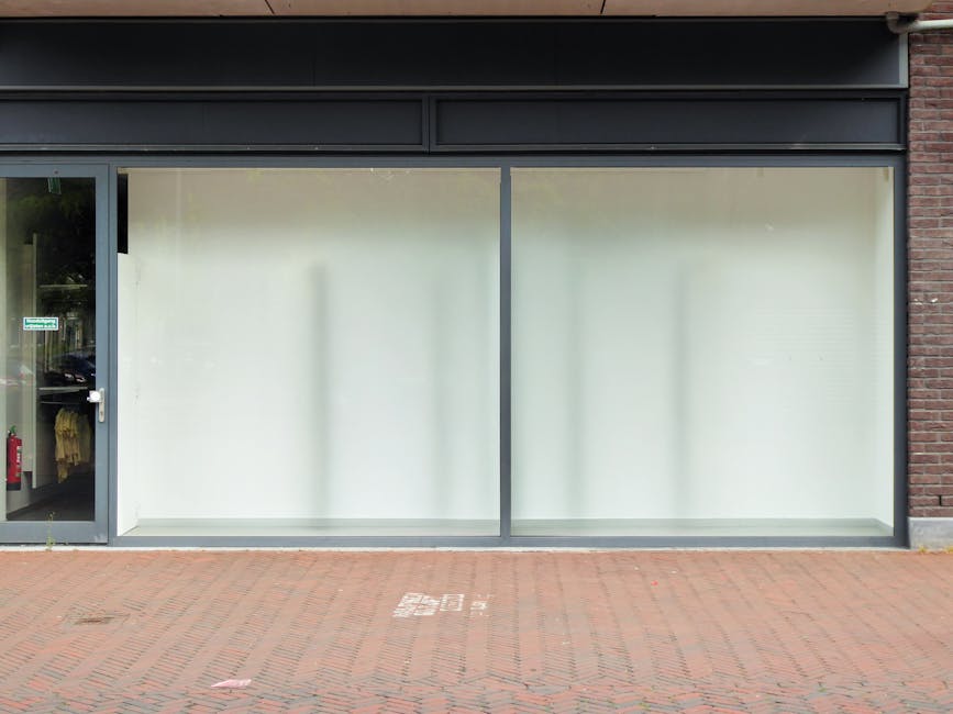The image shows a commercial storefront with large glass windows and a glass door, framed by a metal structure. The windows are frosted or covered with white blinds, giving a clean and tidy appearance